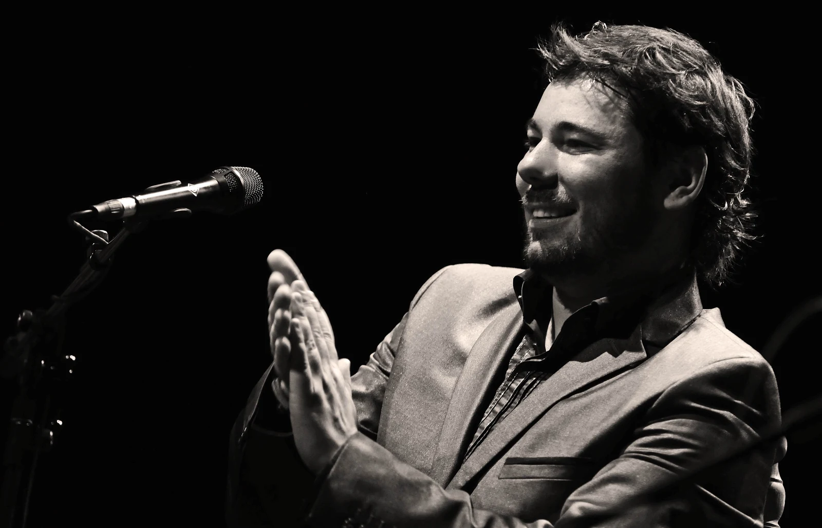 Black-and-white photograph of Miguel Poveda singing into a microphone, with his hands raised near his chest.