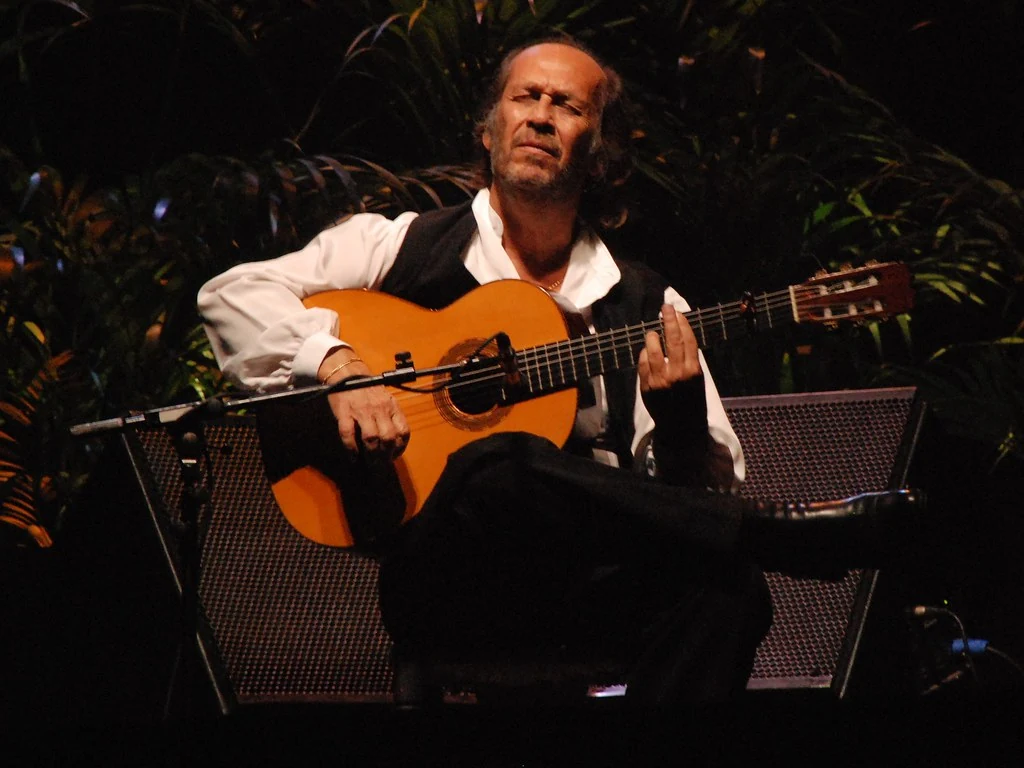 Paco de Lucía seated on stage, playing a flamenco guitar in a white shirt and dark waistcoat.