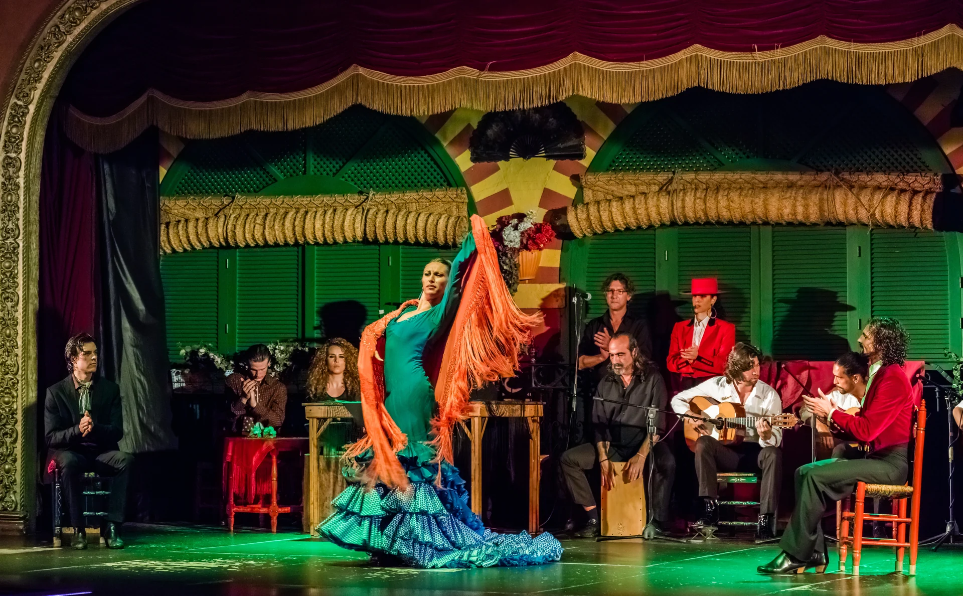 A dancer in a turquoise dress raises an orange shawl, accompanied by musicians at El Palacio Andaluz in Seville.