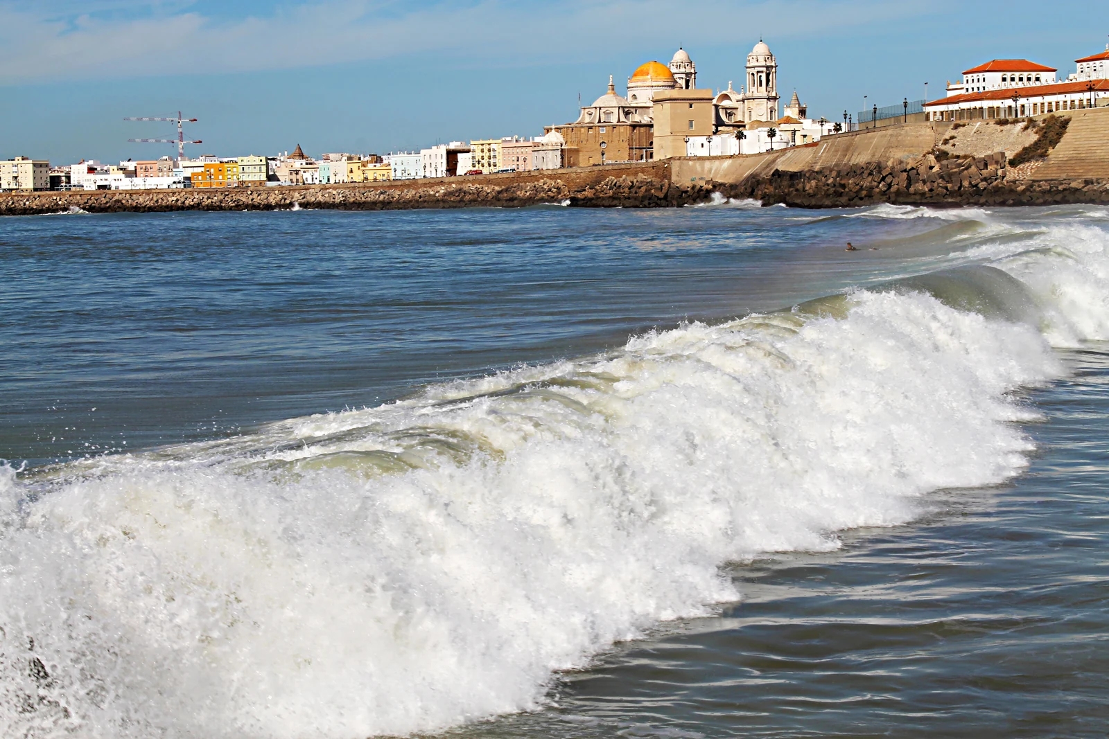 Olas frente al paseo de Campo del Sur, con la cúpula dorada y las torres de la catedral de Cádiz al fondo.