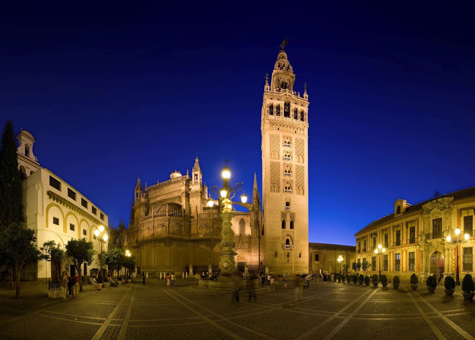 Seville Cathedral and the Giralda illuminated beside Plaza Virgen de los Reyes at blue hour.
